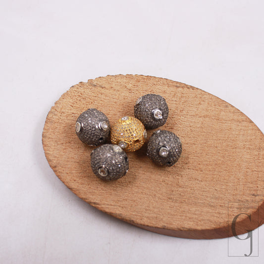 A selection of 13mm pave diamond beads with 925 sterling silver handmade finish, displayed on a wooden surface. One of the beads features a prominent golden hue with a rose cut design, surrounded by smaller gray beads with a similar pave diamond design.