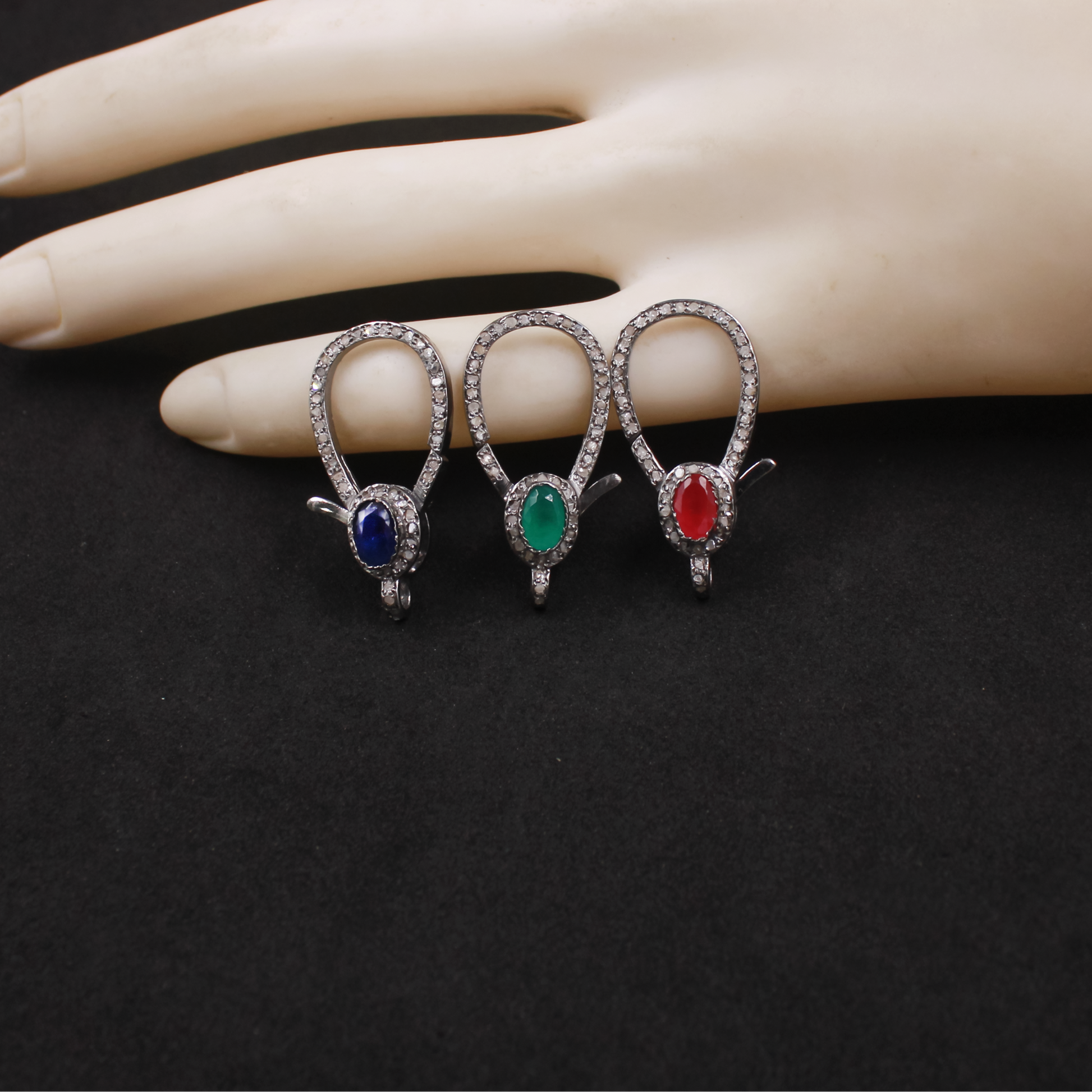 Three heart-shaped jewelry pieces with colored stones on a mannequin hand against a black background