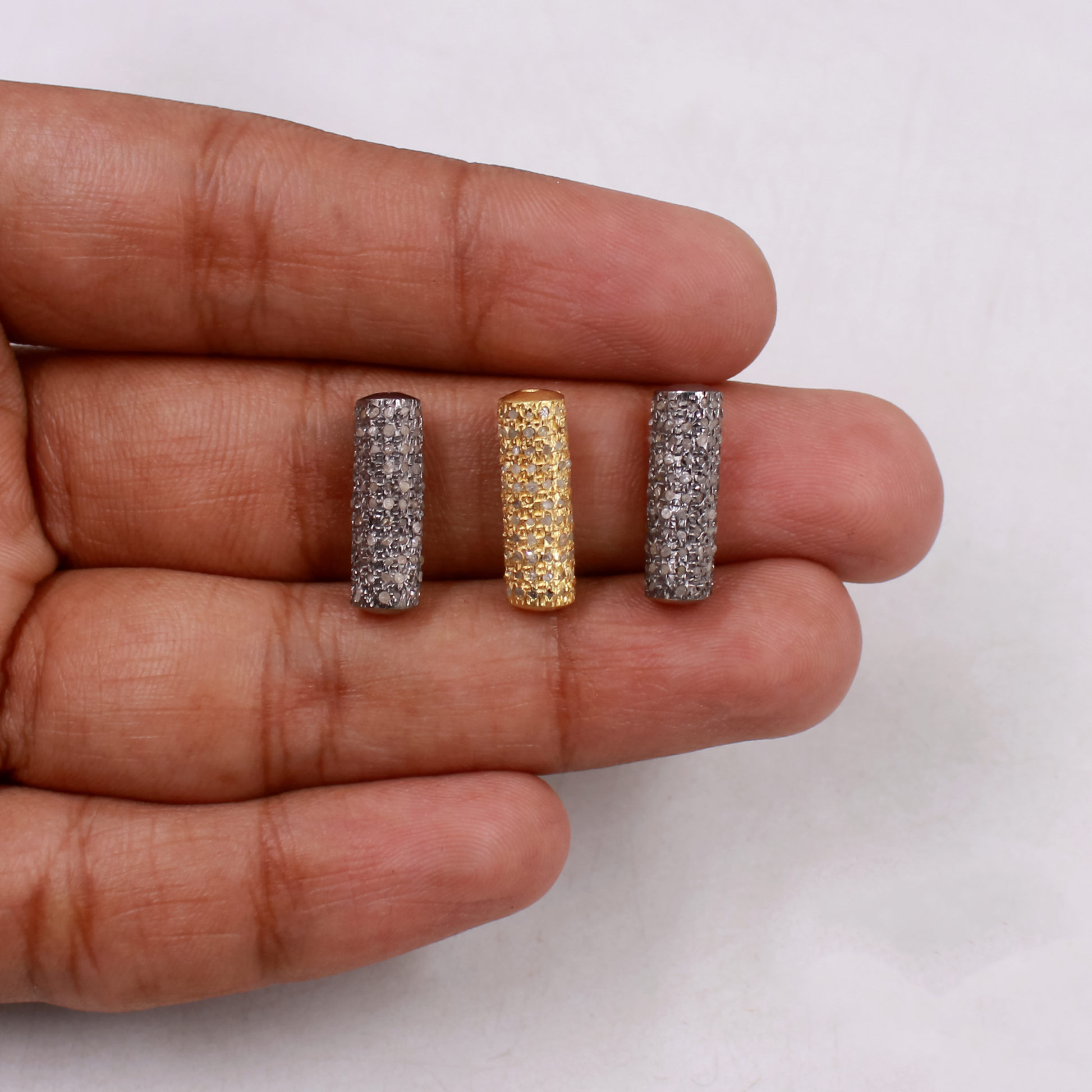 Three cylindrical jewelry pieces in silver, gold, and another silver color held between fingers on a white background.