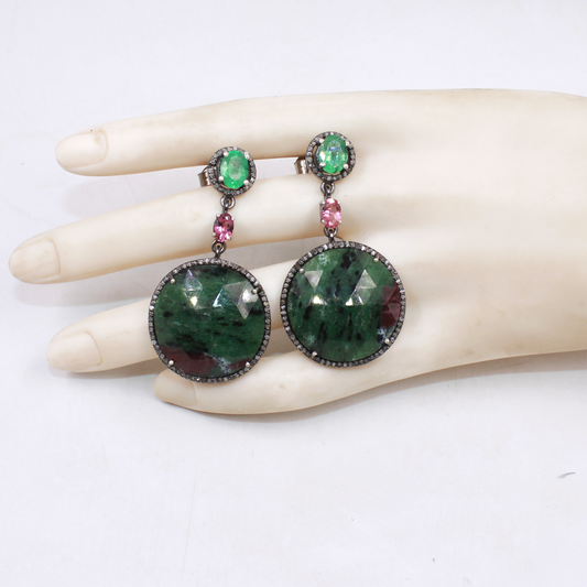 Green gemstone earrings with pink accents on a mannequin hand against a white background
