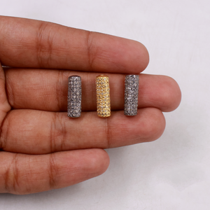 Three cylindrical jewelry pieces in silver, gold, and another silver color held between fingers on a white background.