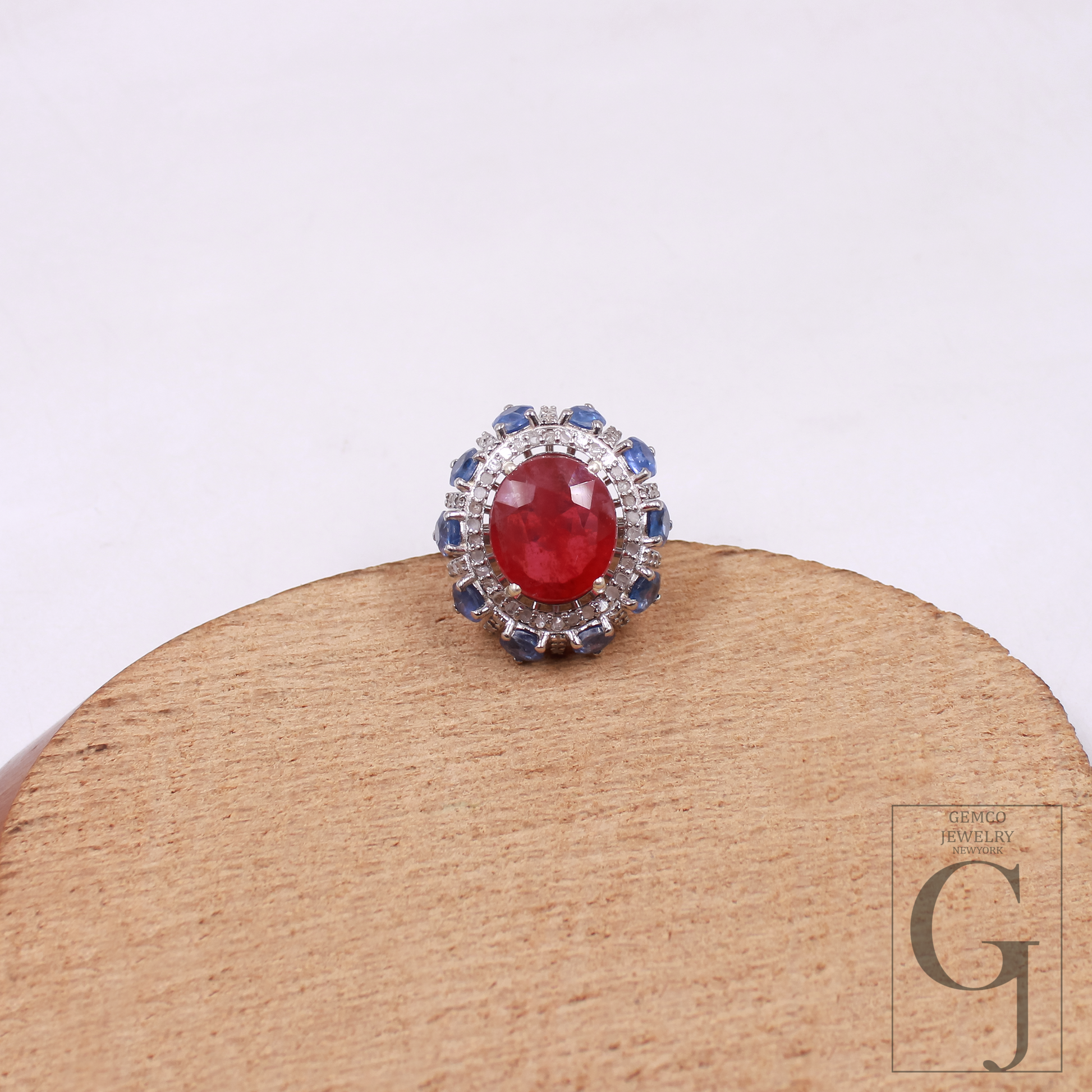Ring with a red gemstone and blue accents on a wooden stand with a white background