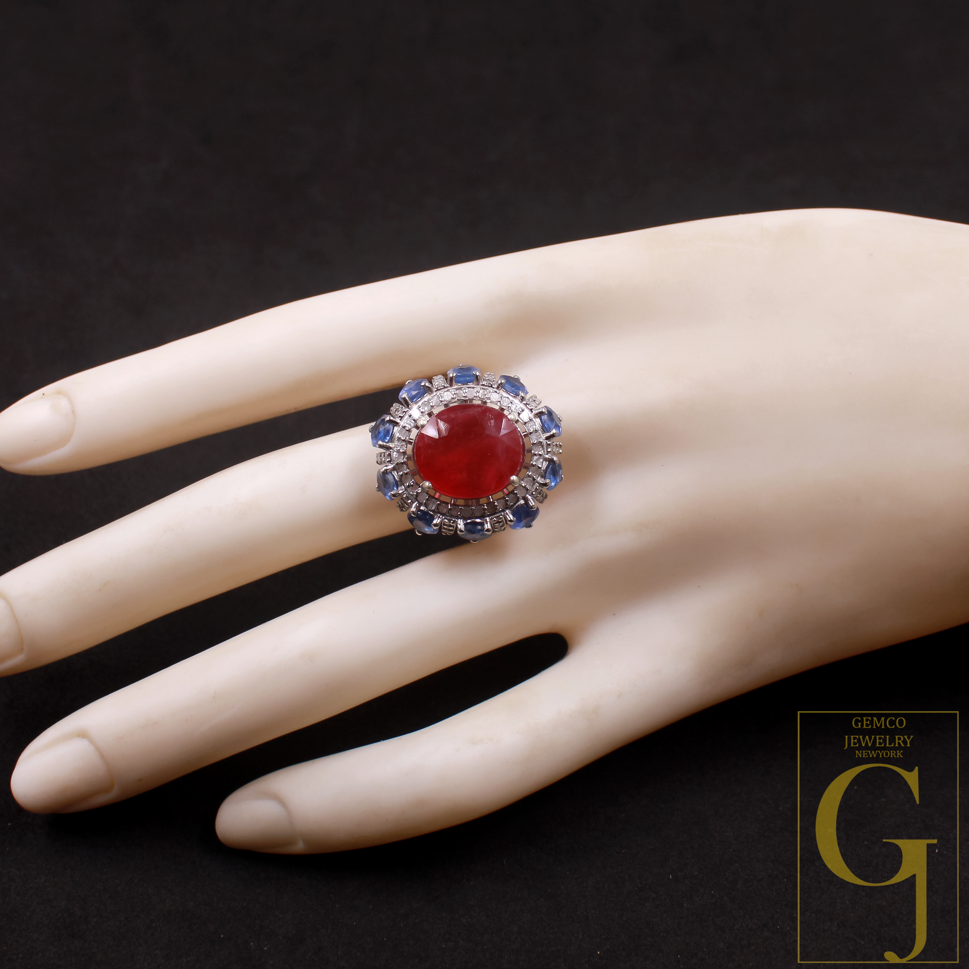 Ring with a red gemstone on a mannequin hand against a dark background, featuring the Gemco Jewelry logo.