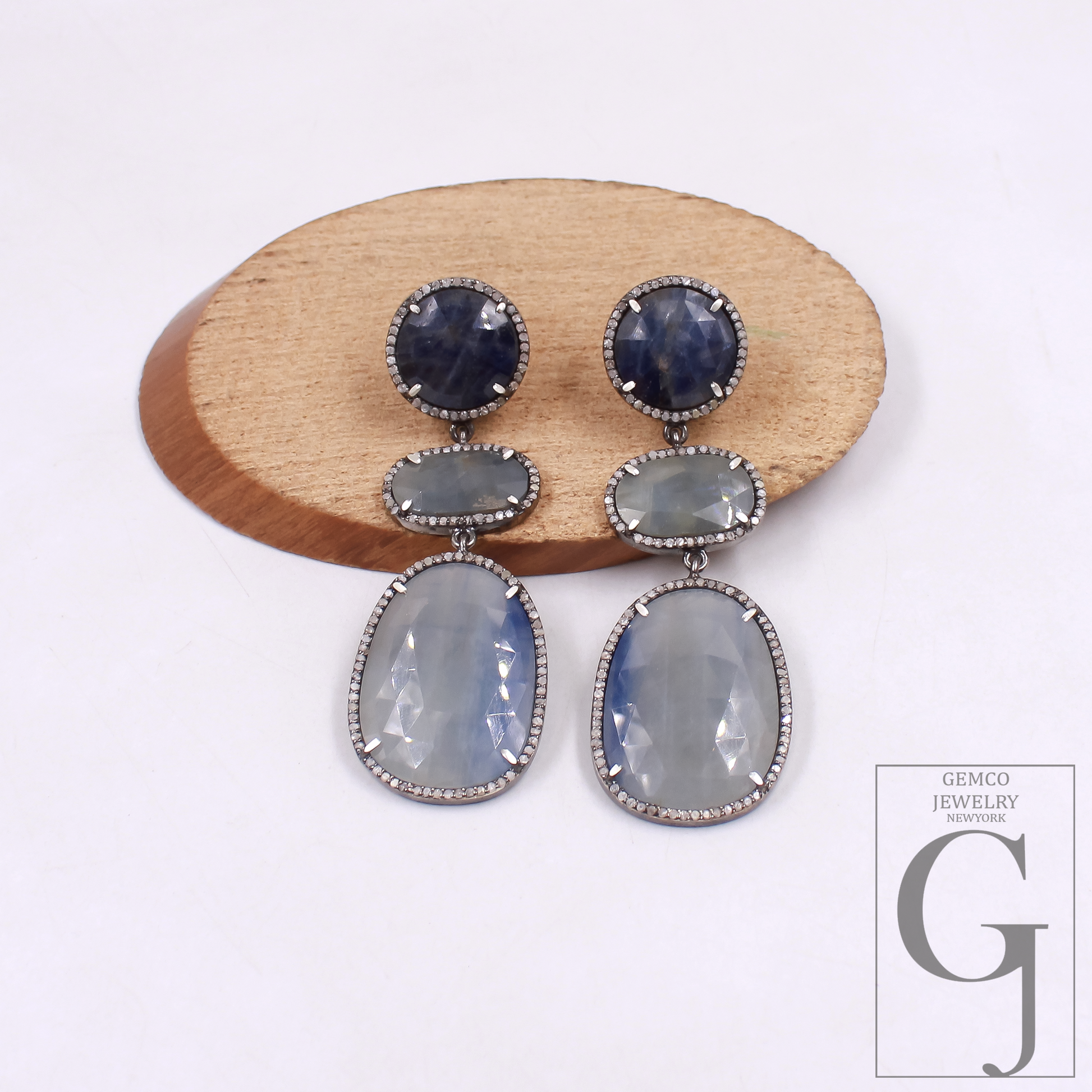 Pair of earrings with gemstones on a wooden stand against a white background, featuring the Gemco Jewelry logo.