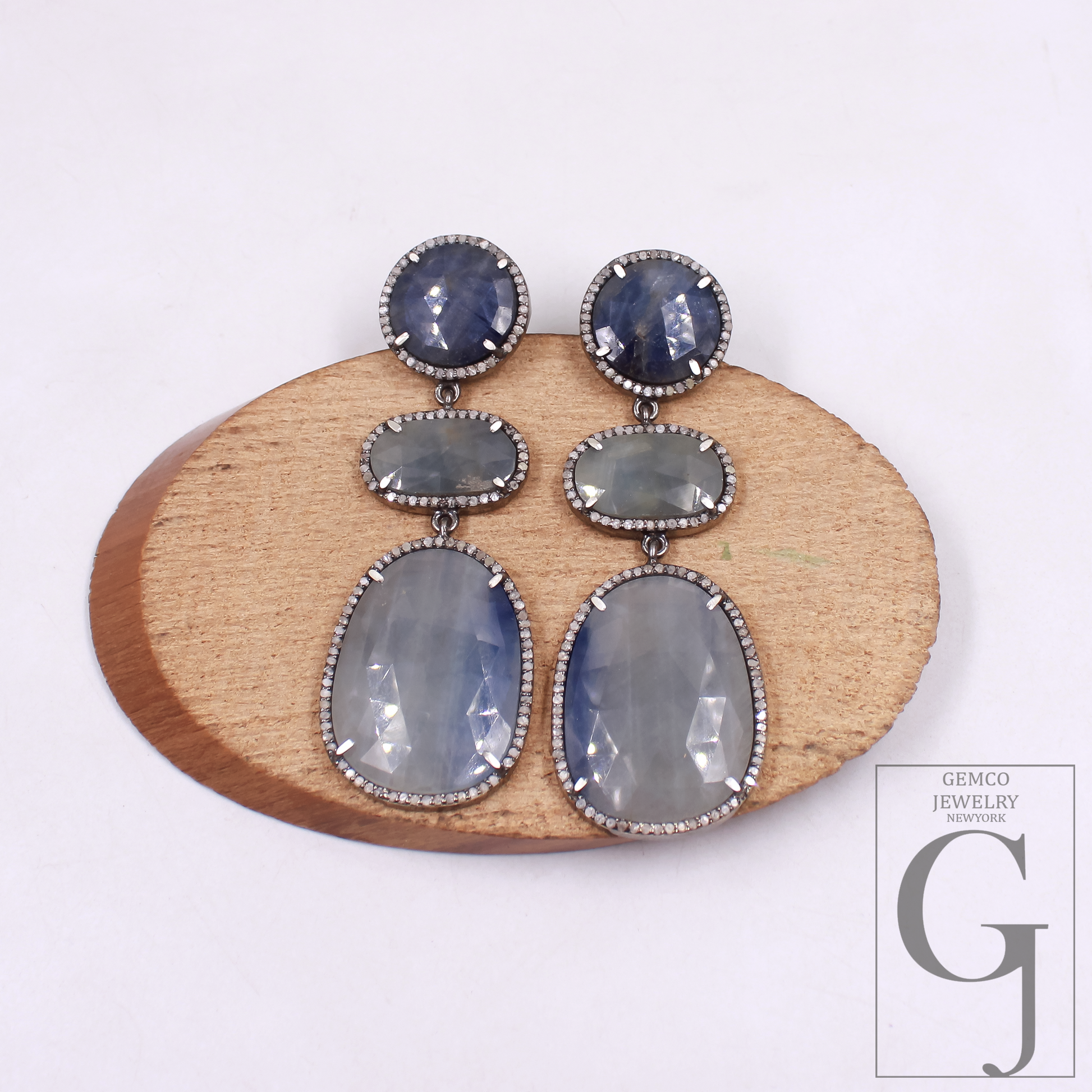 Pair of earrings with gemstones on a wooden stand, brand 'Gemco Jewelry' visible.