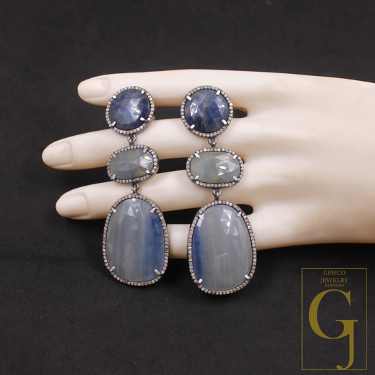 Pair of dangling earrings with blue stones on a mannequin hand against a dark background, featuring the Gemco Jewelry logo.