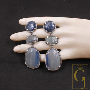 Pair of dangling earrings with blue stones on a mannequin hand against a dark background, featuring the Gemco Jewelry logo.