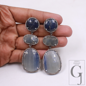Pair of silver earrings with blue and gray stones held in a hand against a white background.