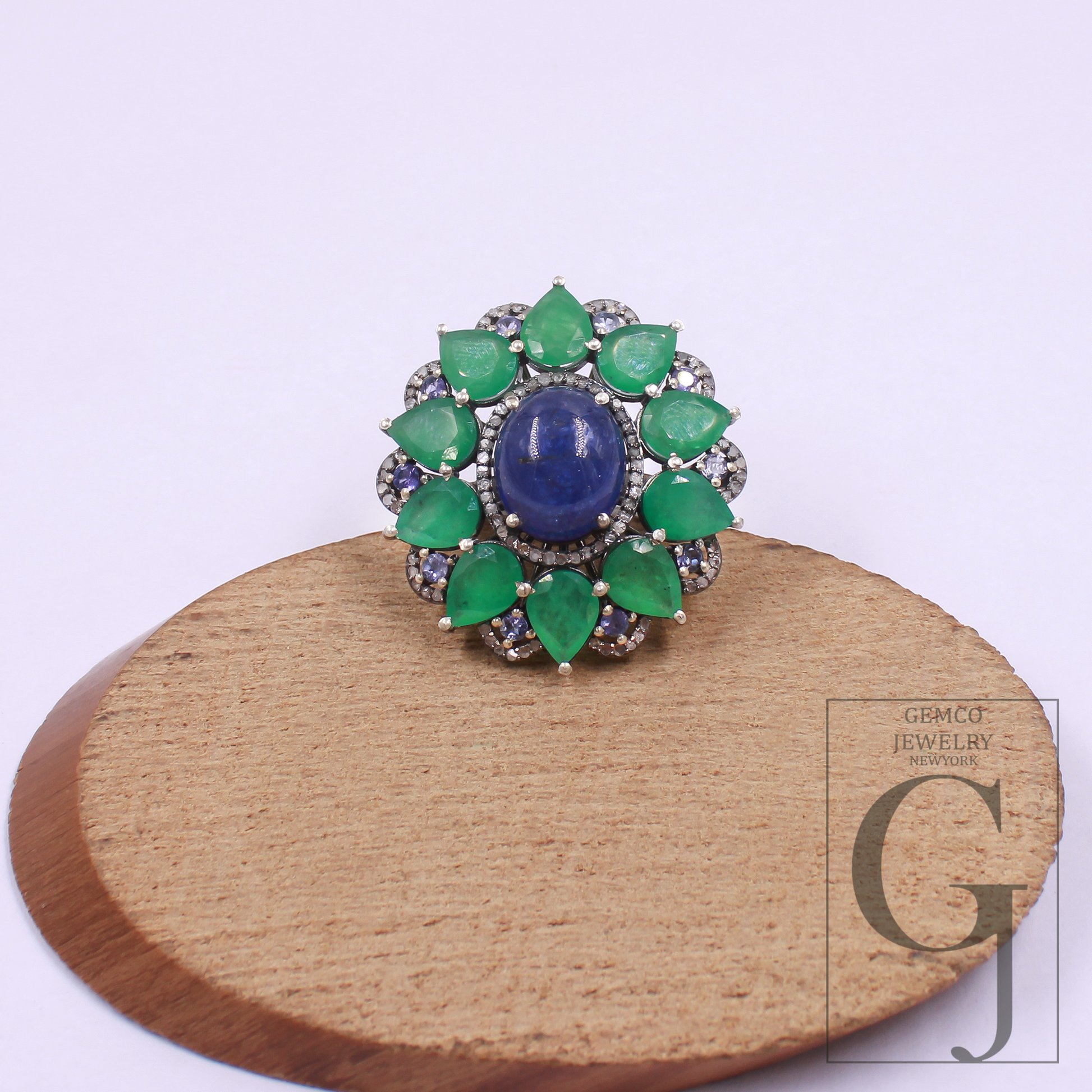 Jewelry ring with green gemstones and a blue center on a wooden stand with 'Gemco Jewelry' branding.
