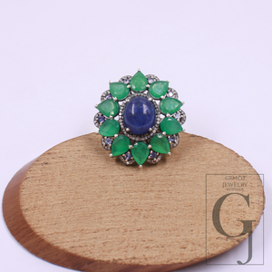 Jewelry ring with green gemstones and a blue center on a wooden stand with 'Gemco Jewelry' branding.