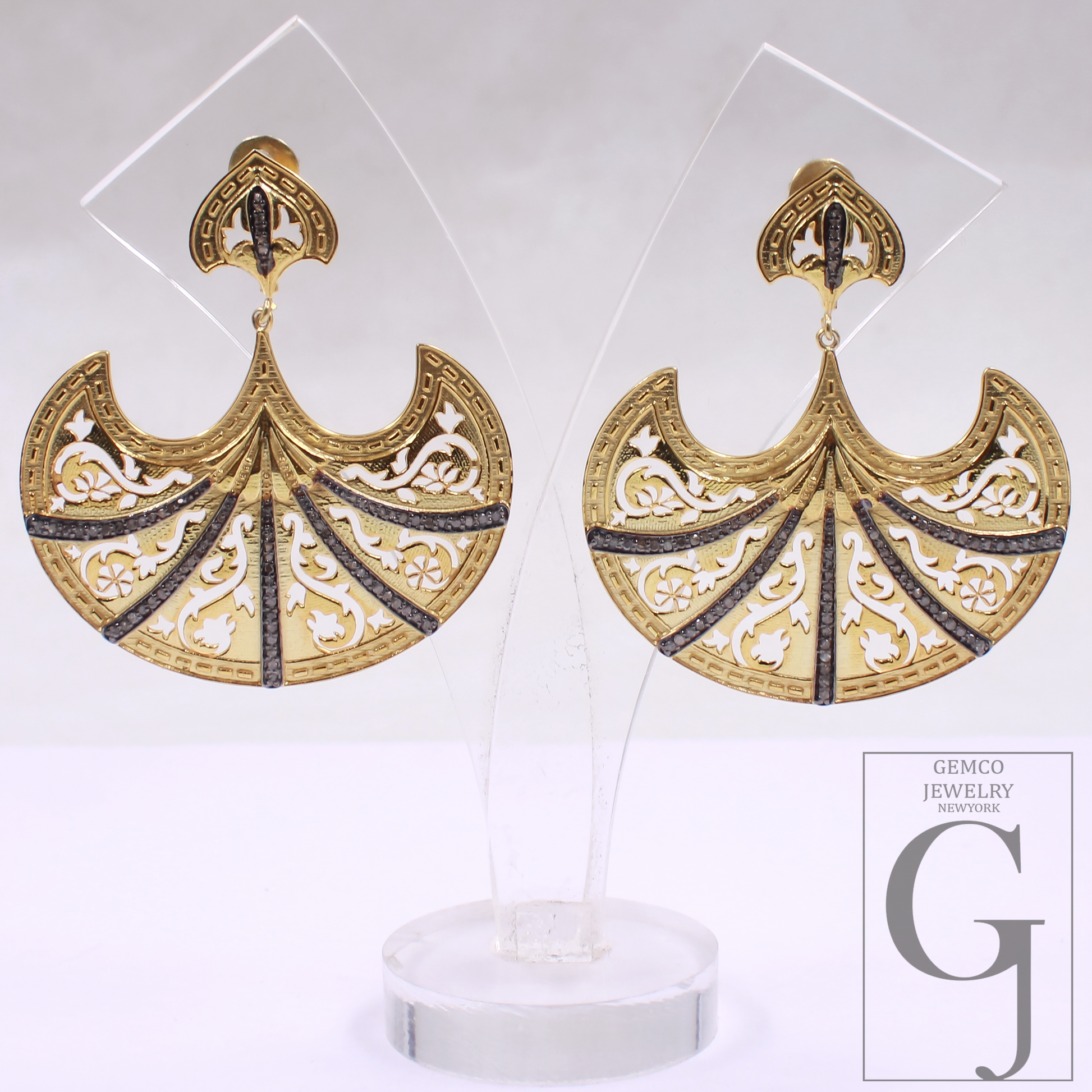 Gold earrings with intricate designs on a clear stand, branded 'Gemco Jewelry'.