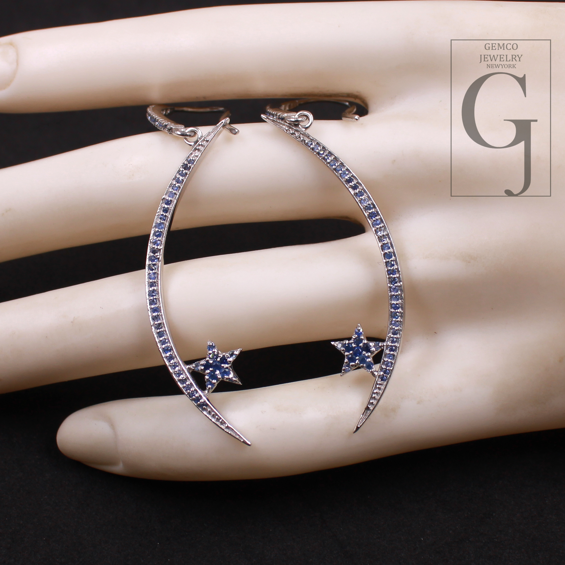 Silver hoop earrings with blue gemstones on a mannequin hand, featuring Gemco Jewelry logo.