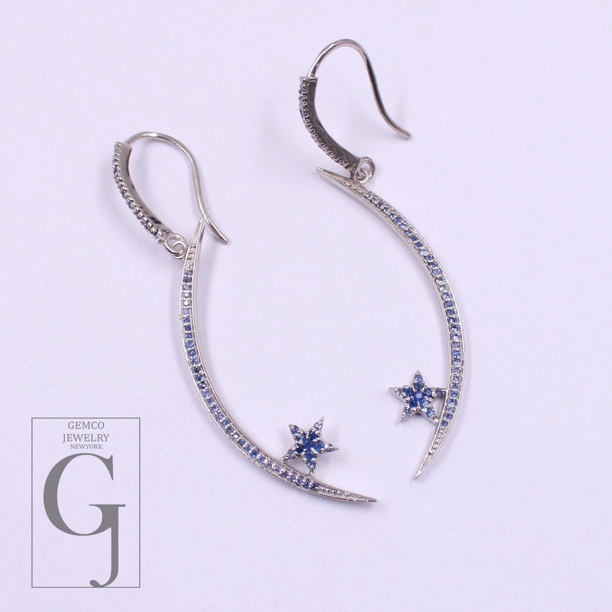 Pair of silver earrings with star and crescent moon designs on a light purple background, featuring Gemco Jewelry New York logo.