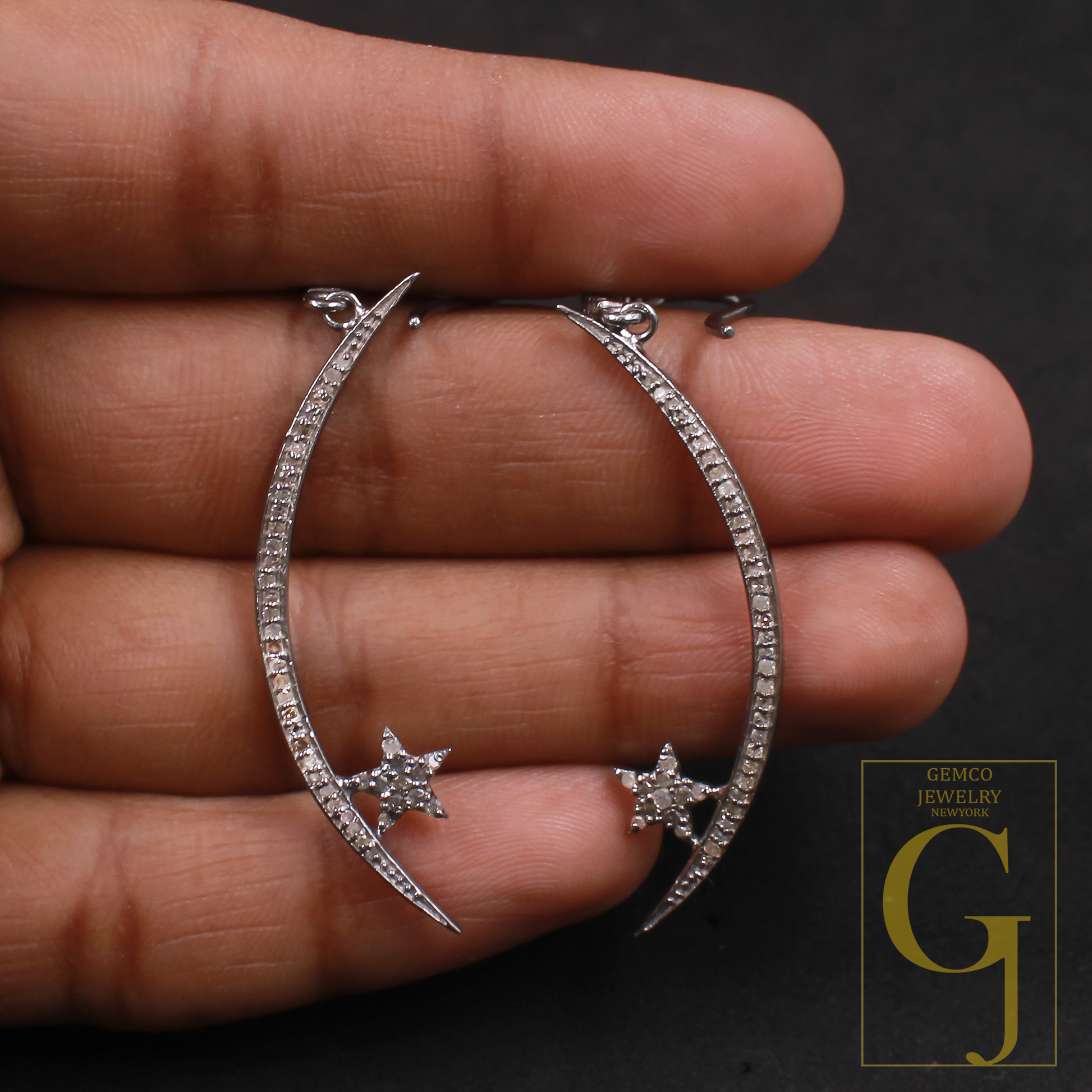 Silver hoop earrings with star designs held between fingers against a dark background, featuring Gemco Jewelry logo.